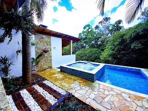 A pool area with a stone and wooden deck, a hot tub, and lush greenery.