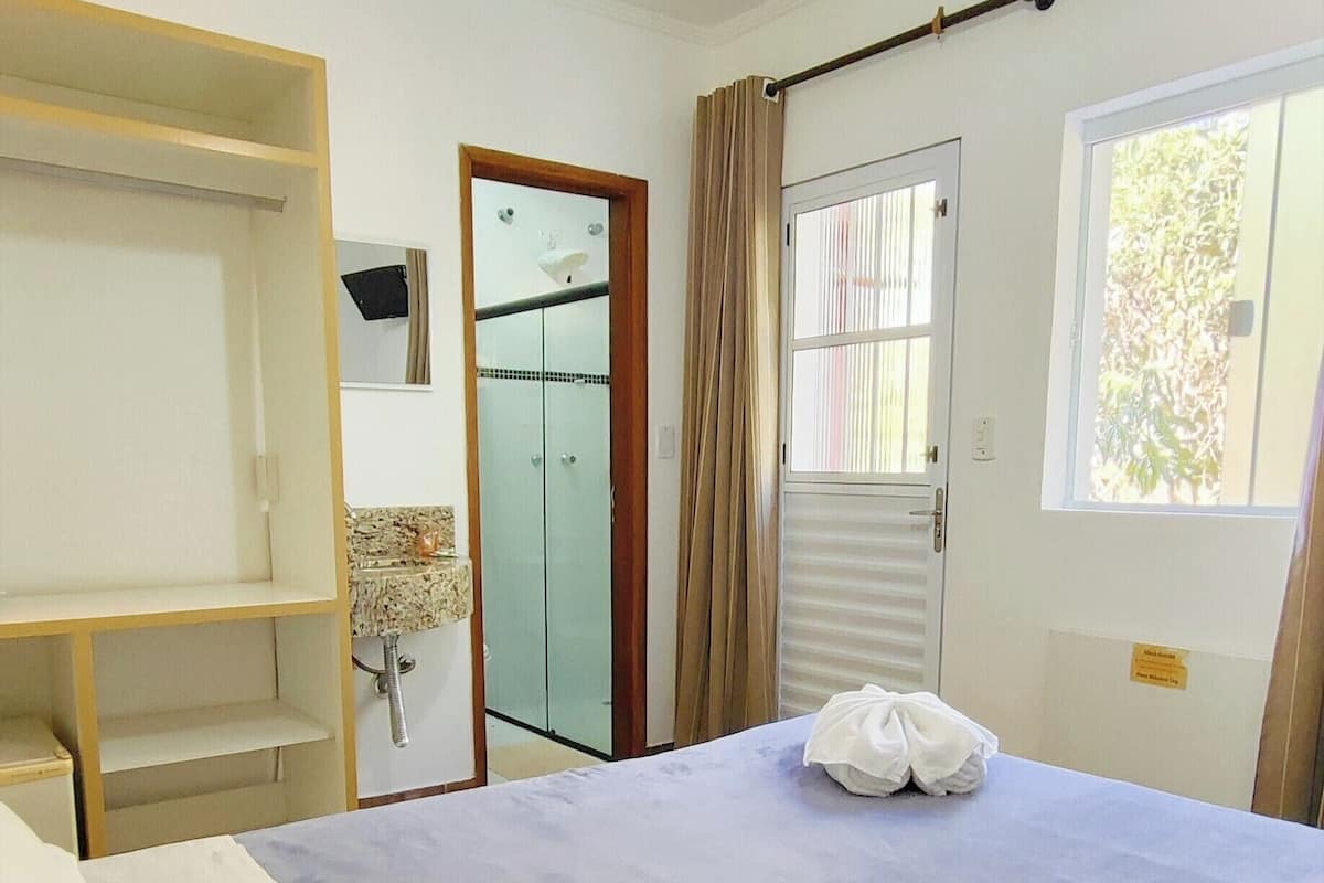 Habitación de hotel con cama, armario, baño con puerta de vidrio y ventana con cortinas.
