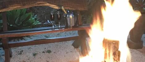 A campfire with logs and burning embers, surrounded by a wooden bench and a metal pot.
