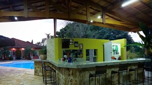A bar area with a stone counter and bar stools, a pool in the background, and a building with a red roof. - Pousada Della Vegas (Serra Negra)