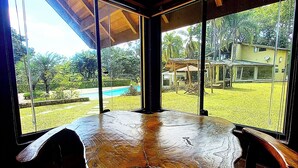A wooden table with four chairs in a room with large windows overlooking a backyard with a pool and a house. - Pousada Della Vegas (Serra Negra)