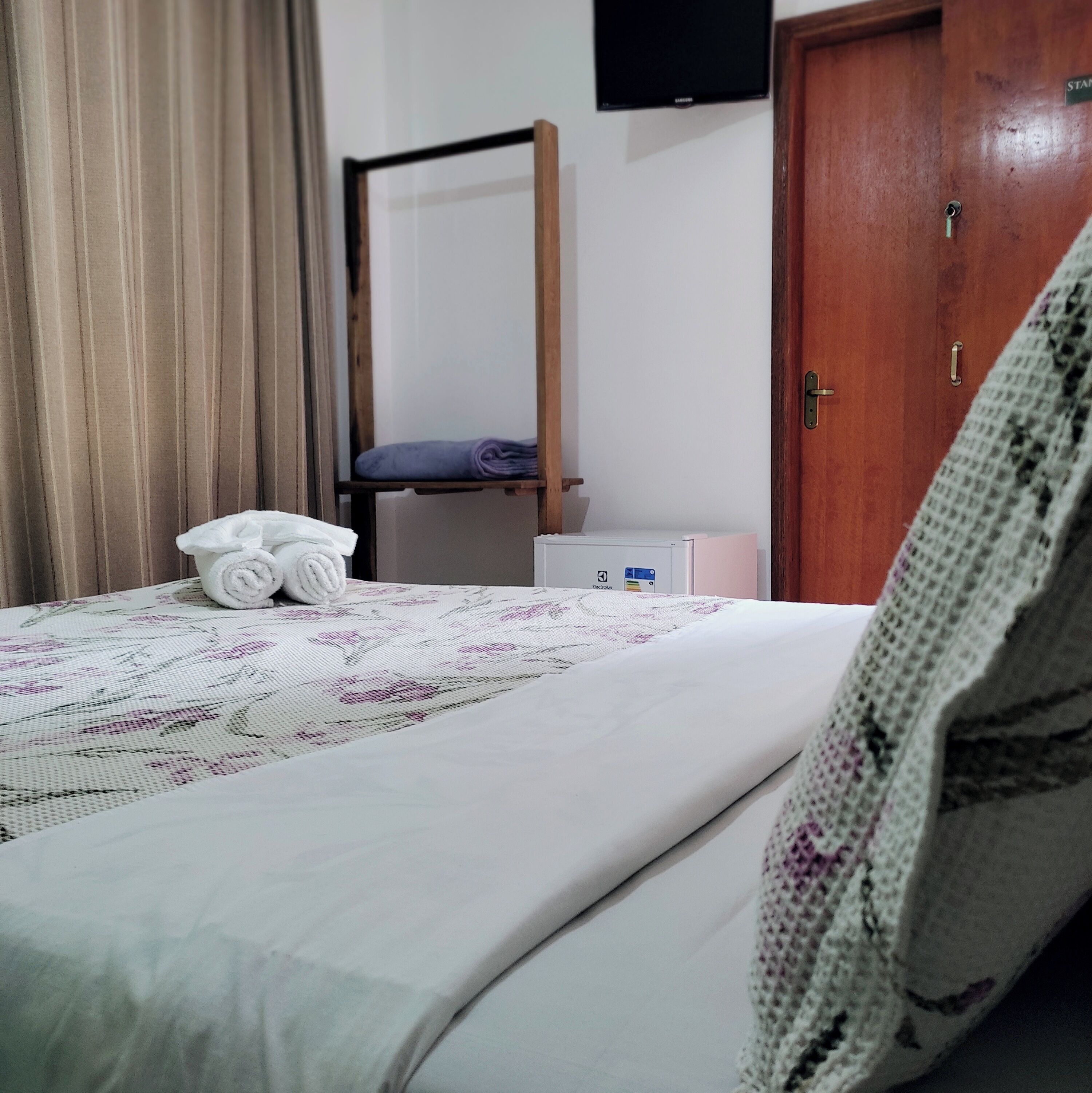 A neatly made bed with a floral-patterned bedspread, a rolled white towel, and a folded purple towel on the bed.