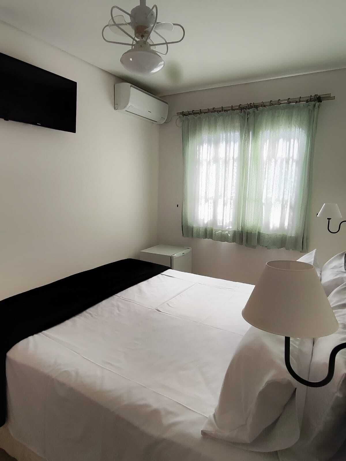 A hotel room with a bed, a bedside table, a lamp, an air conditioning unit, and a window with curtains.