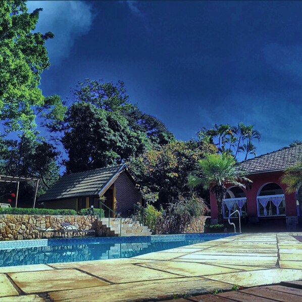 A pool area with a stone deck, a small building with a tiled roof, and a large tree in the background.