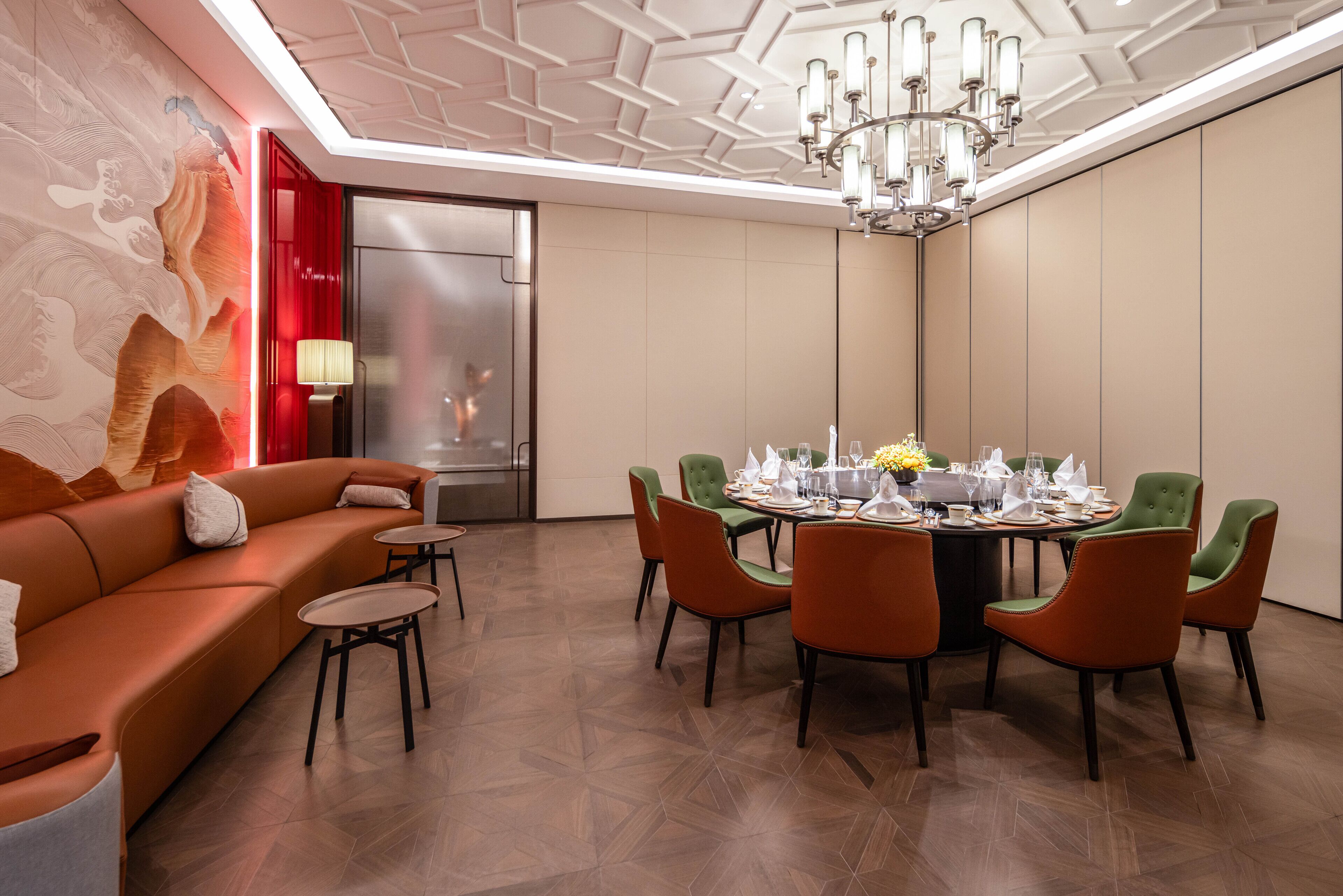 Un espace repas moderne avec une table ronde pour six personnes, entourée de chaises orange, et un canapé devant un mur d’accent rouge.