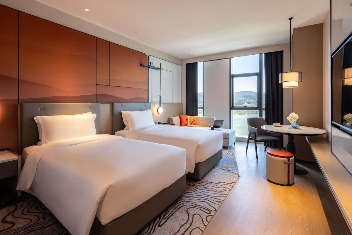 Habitación de hotel con dos camas, una mesita con una silla, vistas al exterior y un ventanal grande.