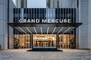 L’entrée de l’hôtel Grand Mercure se distingue par sa façade moderne, ses grandes fenêtres en verre et un panneau lumineux bien visible.