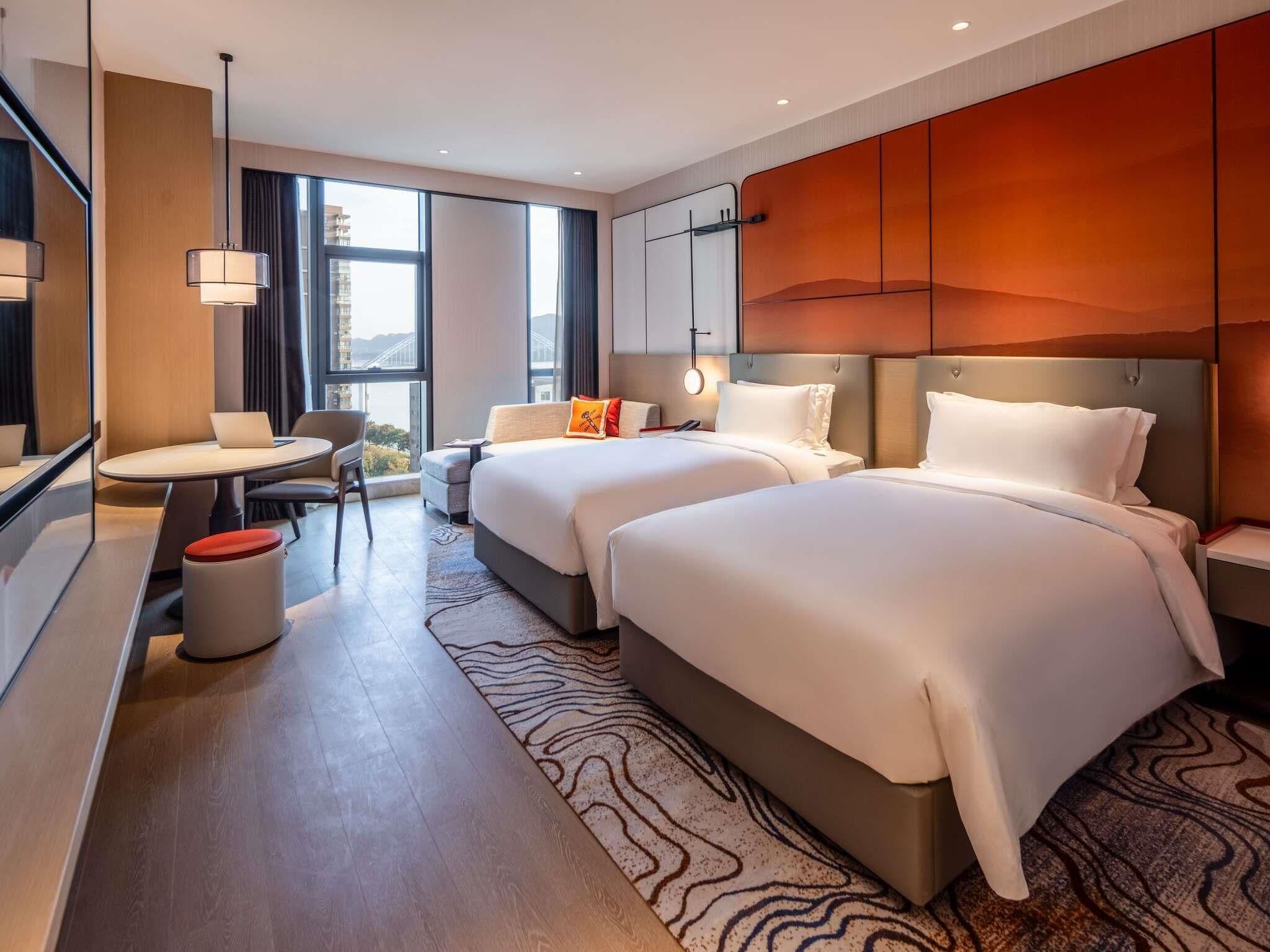 A modern hotel room with two beds, a desk, a chair, and a view of a bridge.