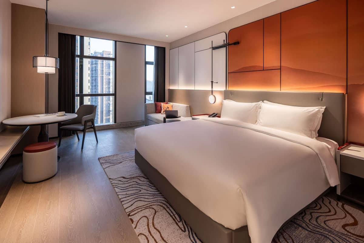 Habitación de hotel moderna con una cama grande, un escritorio, una silla y vistas de la ciudad a través de amplias ventanas.