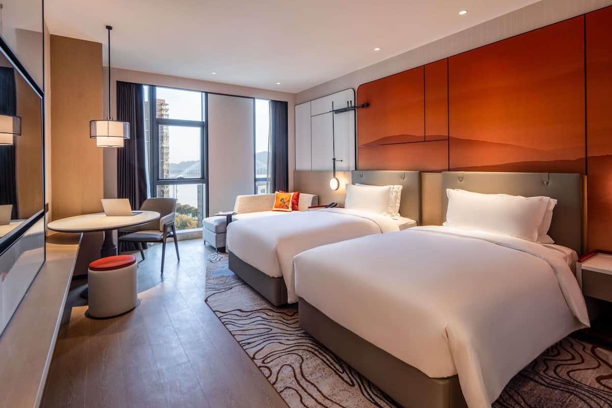 Habitación de hotel con dos camas, un escritorio con silla, una mesita y vistas a un puente.