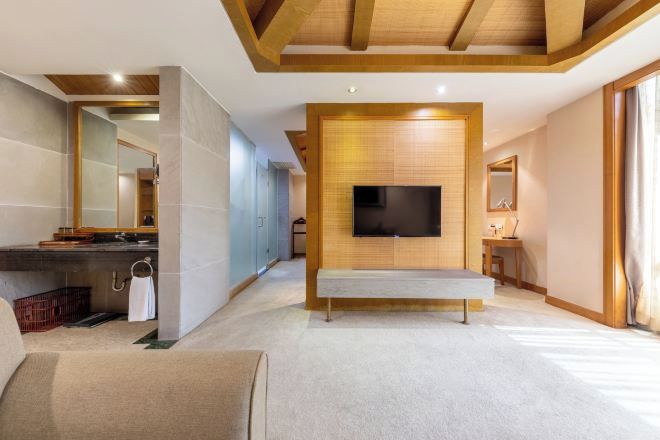 A modern hotel room with a large flat-screen TV mounted on a wooden panel, a bench in the center, and a large mirror on the left wall.