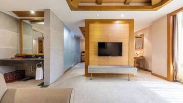 A modern hotel room with a large flat-screen TV mounted on a wooden panel, a bench in the center, and a large mirror on the left wall.