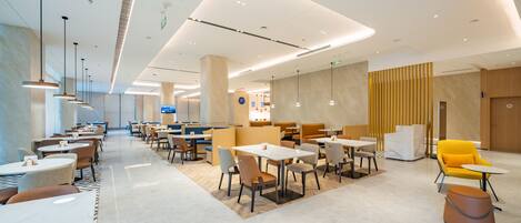 A modern hotel lobby with multiple seating areas, including tables and chairs, and a central area with a yellow chair and ottoman.