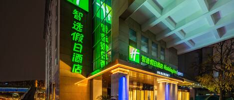 A hotel entrance with illuminated signage, a staircase leading to the entrance, and outdoor seating with potted plants.