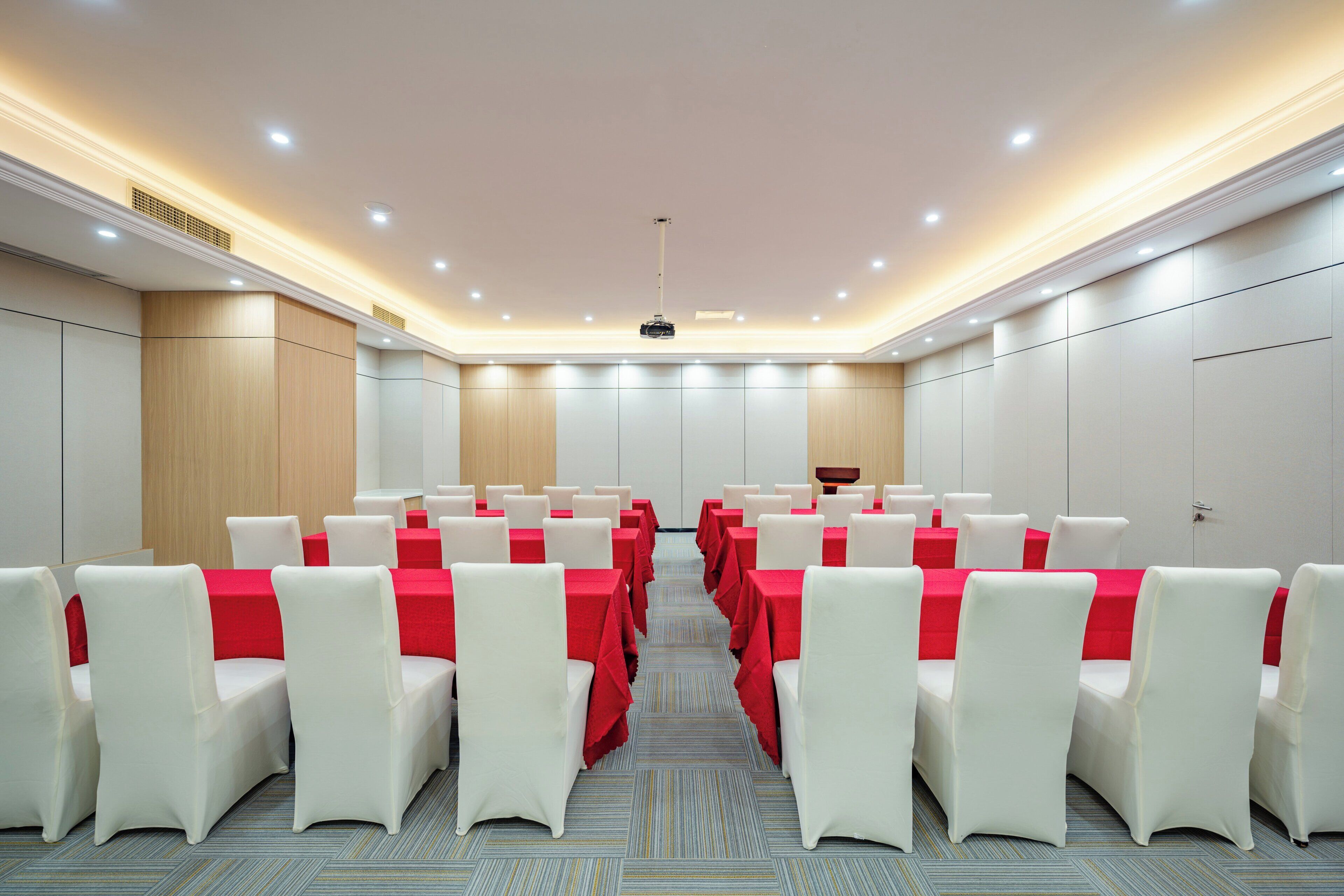 A conference room with rows of white chairs and red tablecloths, equipped with ceiling lights and a projector.