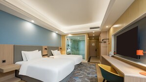 Ein Hotelzimmer mit zwei Betten, einem Schreibtisch, einem Stuhl, einem Fernseher und einem durch eine Glastür einsehbaren Badezimmer.