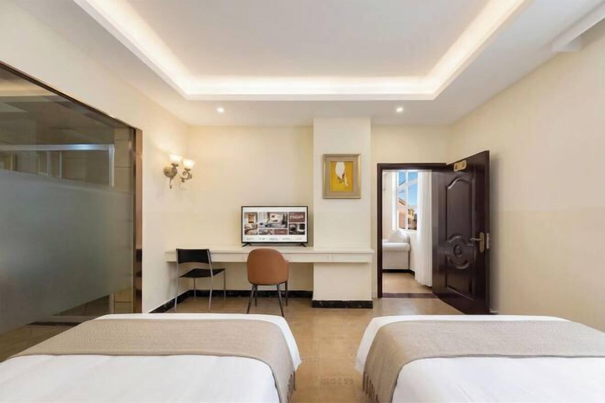 Habitación de hotel con dos camas, un escritorio con una silla, un cuadro en la pared y una puerta que da a otra habitación.
