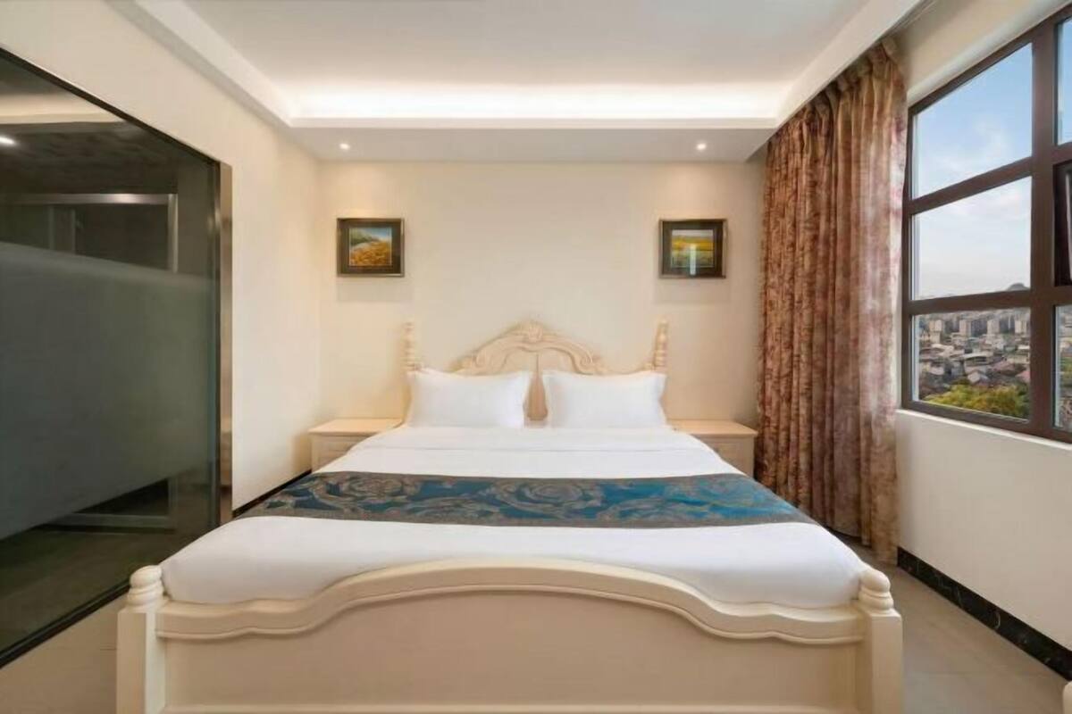 Habitación de hotel con una cama grande, una mesita de noche, una ventana con cortinas y vistas a la ciudad.