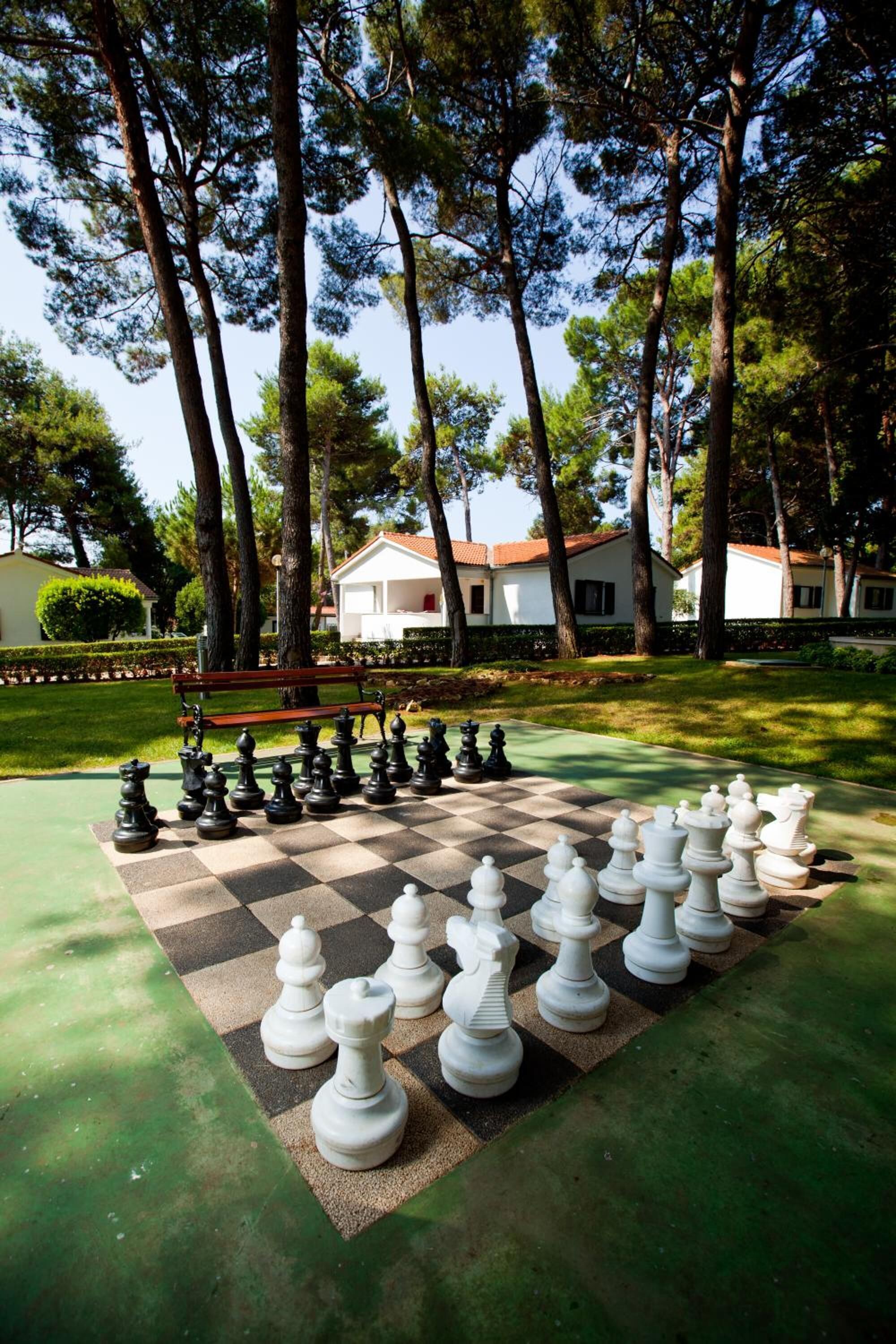 Un tablero de ajedrez grande, al aire libre, dispuesto en un parque con árboles y un edificio blanco al fondo.