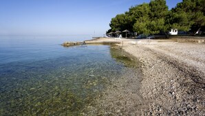 A pebble beach with clear water, a small pavilion, and a white building surrounded by trees. - Villa Galijot Plava Laguna (Porec)