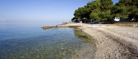 A pebble beach with clear water, a small pavilion, and a white building surrounded by trees.