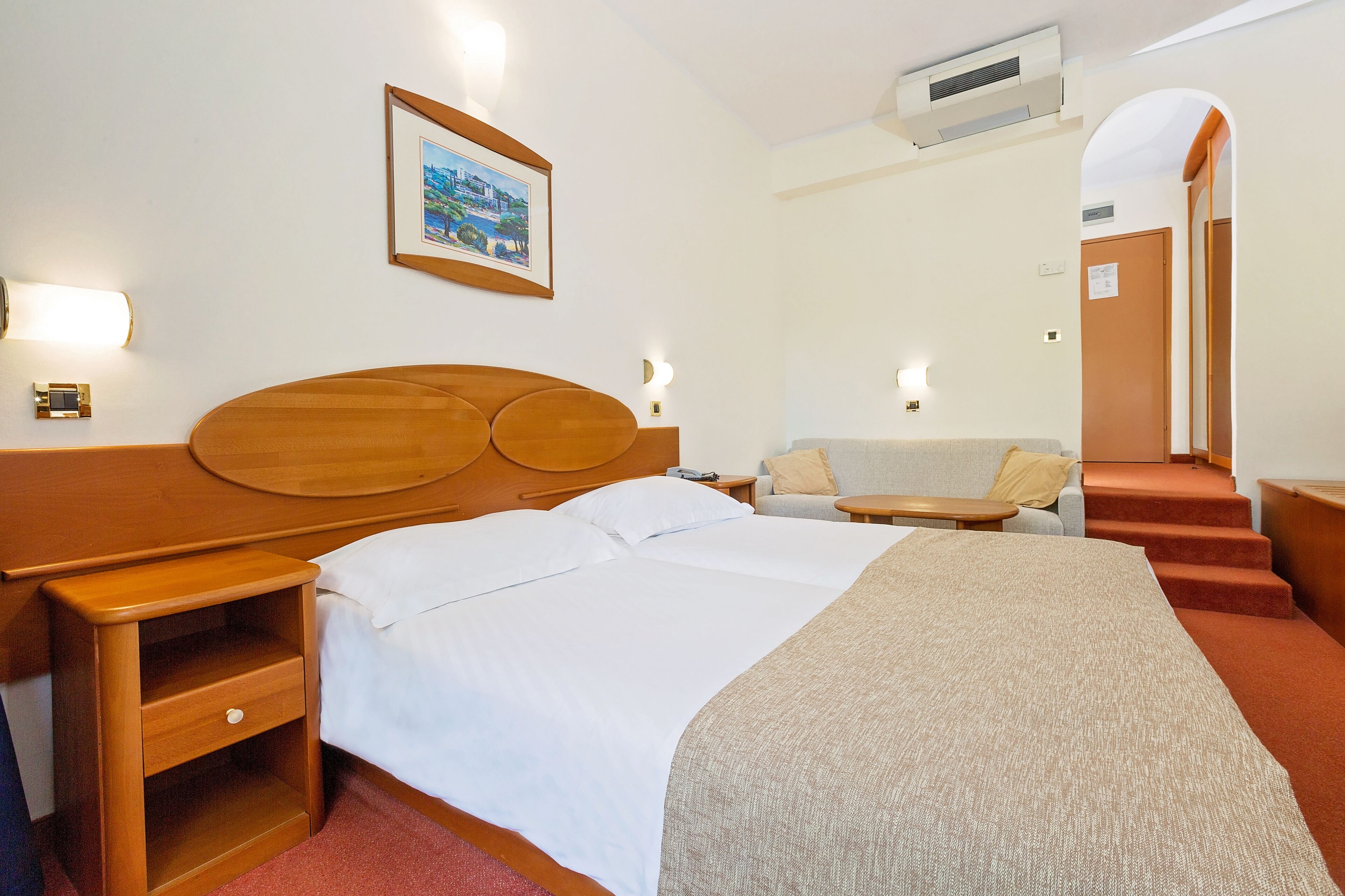 A hotel room with a large bed, a wooden nightstand, a desk, a sofa, and a staircase leading to an upper level.
