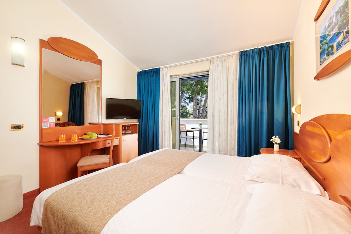 A hotel room with a bed, a desk with a chair, a television, and a balcony with curtains.