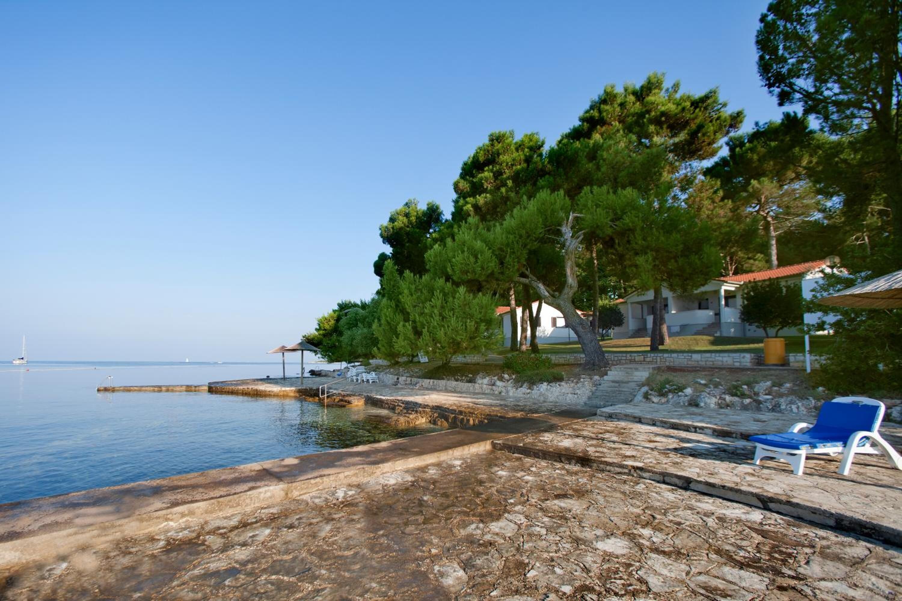 A coastal area with a stone pier extending into calm waters, a row of white lounge chairs, and a row of trees and buildings in the background.