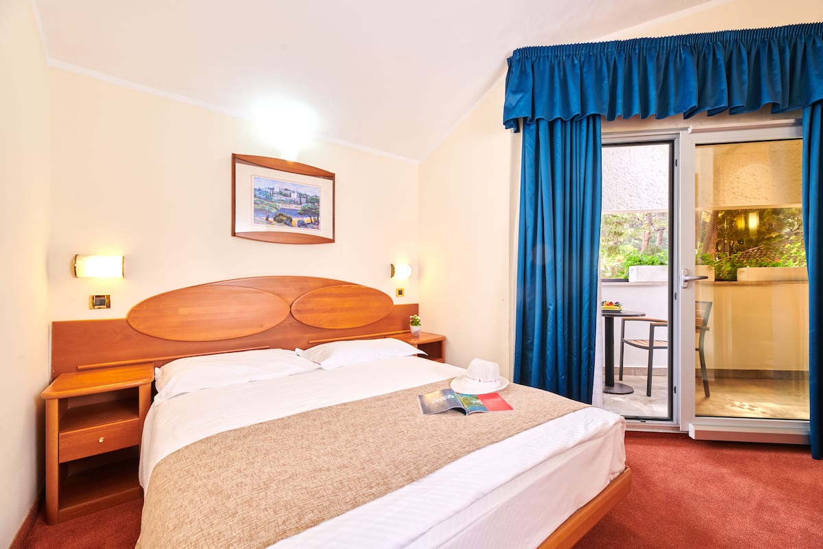 A hotel room with a wooden bed, a nightstand, a framed picture on the wall, and a view of an outdoor area through a sliding glass door.