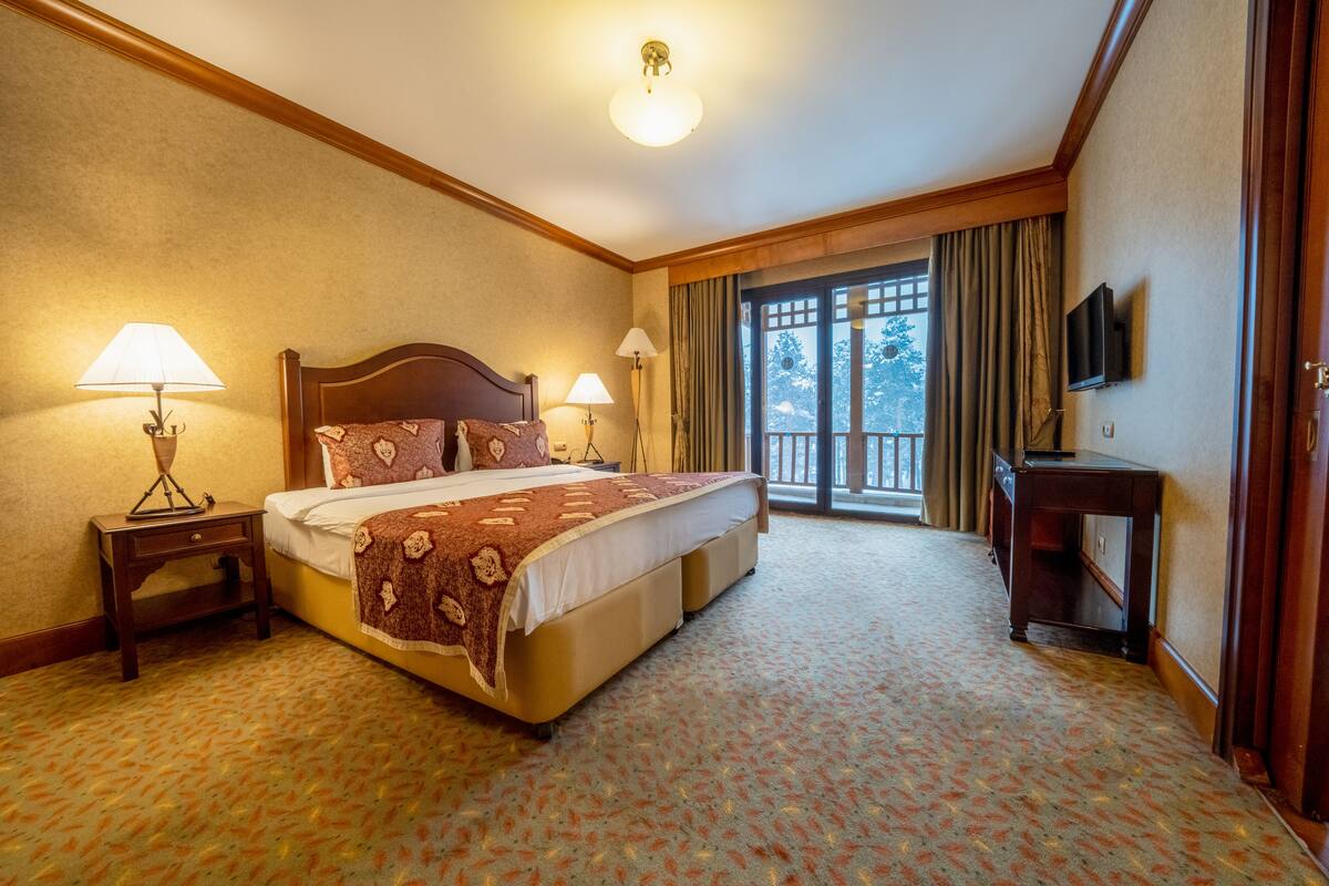 Een hotelkamer met een groot bed, twee bedlampen, een televisie en uitzicht op het omliggende landschap door een raam met gordijnen.