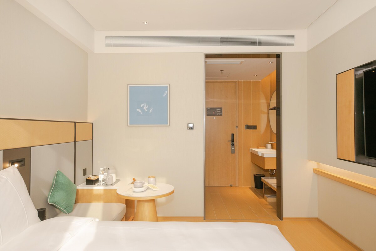 A hotel room with a bed, a small table with amenities, a painting, and a bathroom visible through an open door.