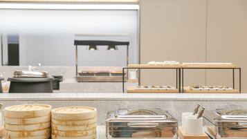 Espaço de buffet com diversos recipientes de comida e uma área de estar ao fundo.