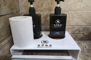 Un ripiano del bagno con un rotolo di carta igienica, due flaconi neri di schiuma da bagno e una scatola bianca con il logo dell'HOTEL JINGHUA.