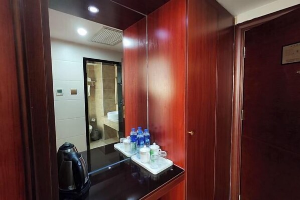 Camera d'albergo con un armadio in legno, un piano di lavoro nero con bollitore e bottiglie d'acqua, e uno specchio che riflette il bagno.