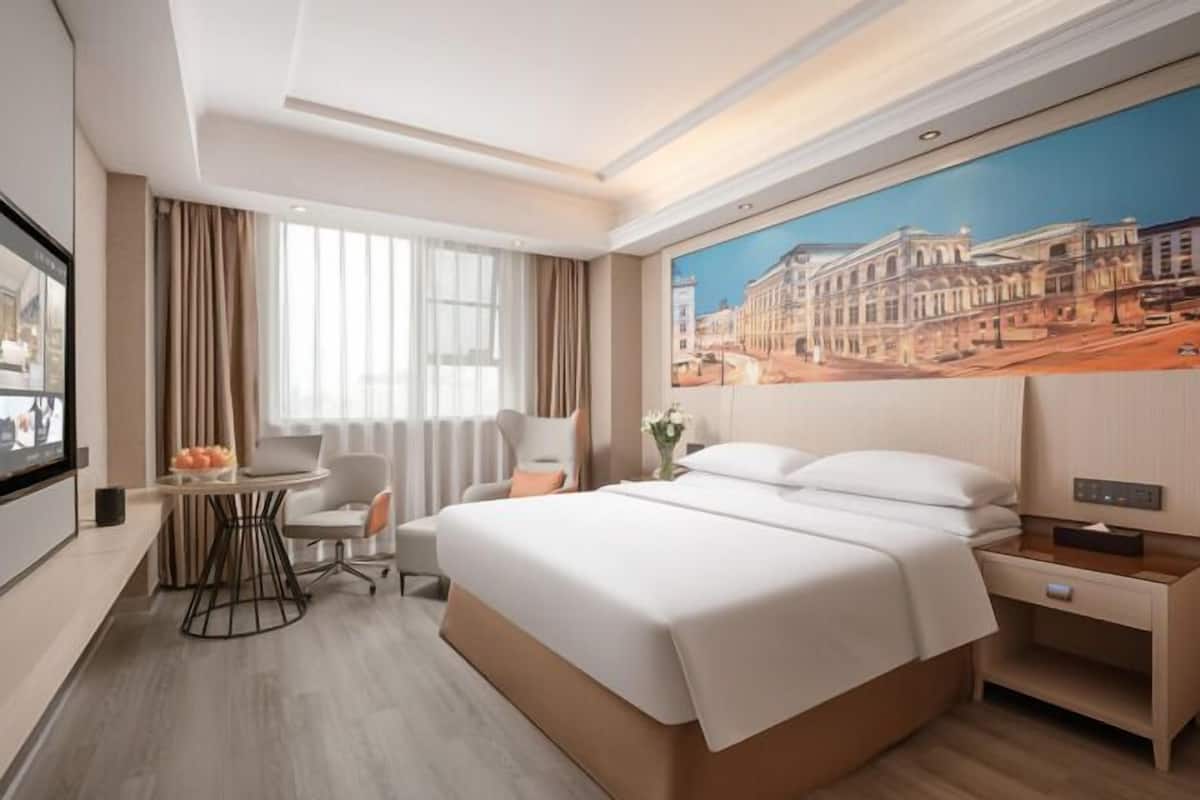 A modern hotel room with a large bed, a desk, a chair, a TV, and a window with curtains.