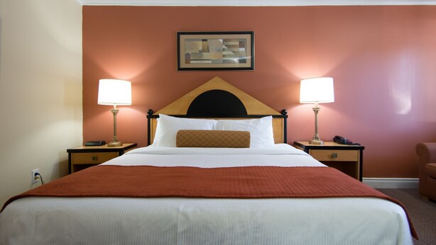Habitación de hotel con una cama grande, dos mesitas de noche con lámparas, una obra enmarcada sobre la cama y un sillón marrón en la esquina.