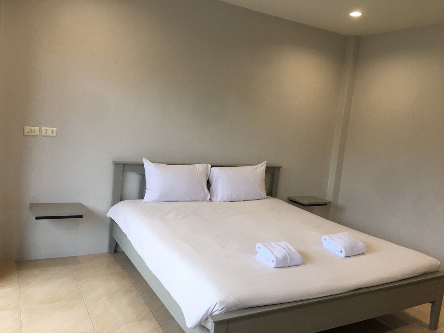Una cama bien hecha con sábanas blancas y dos almohadas, una mesita de noche con una lámpara y un enchufe en la pared.
