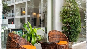 A glass-enclosed outdoor seating area with wicker chairs and a glass table, surrounded by potted plants and a building in the background. - Chaweng terrace (Koh Samui)