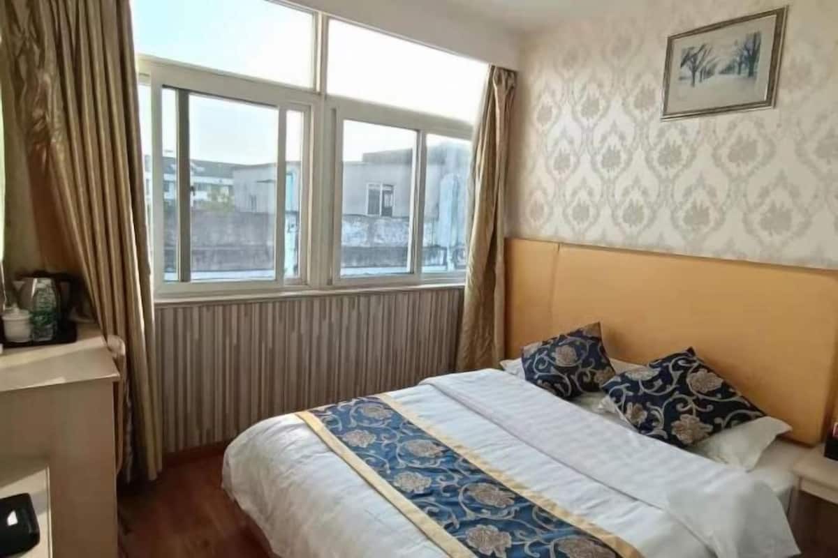 A hotel room with a large window, a bed with patterned bedding, a headboard, and a wall-mounted picture.