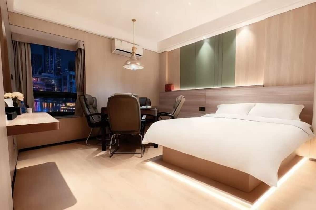 A modern hotel room with a large bed, a desk, two chairs, a view of the cityscape, and a large window.