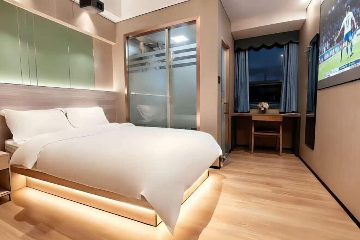 A modern hotel room with a bed, a desk, and a TV screen displaying a sports event.