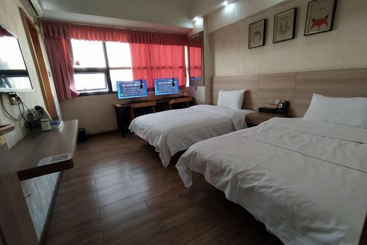 A hotel room with two beds, a desk with two computers, a TV, and a window with red curtains.