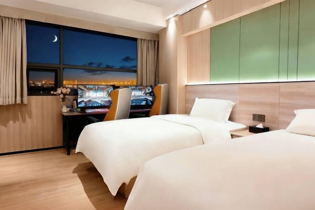 A hotel room with two beds, a desk with a computer, and a view of the cityscape at night.