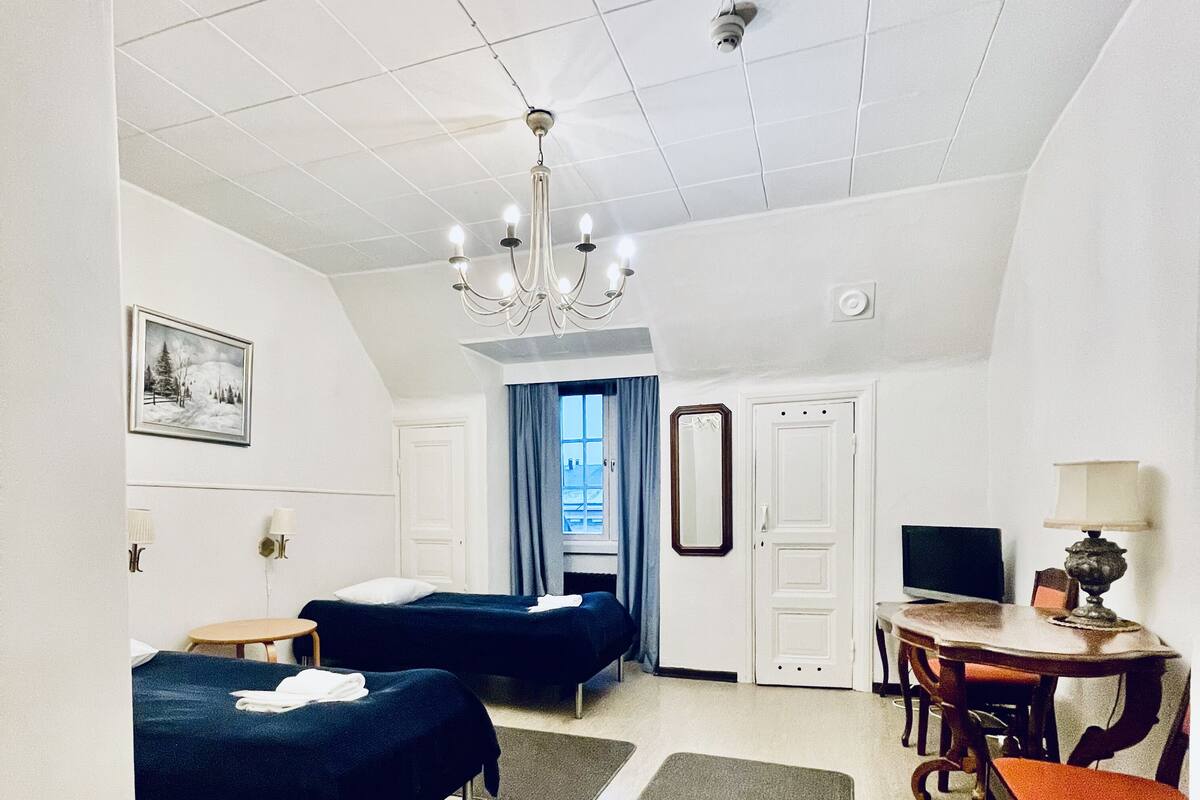 Ein Hotelzimmer mit zwei Betten, einem Schreibtisch, einem Sessel, einem Fernseher, einer Chandelüster, einem Gemälde und einem Fenster mit Vorhängen.