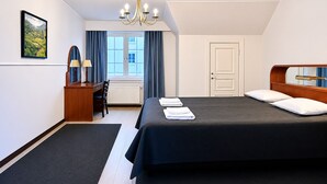 A hotel room with a large bed, a desk with a chair, a desk lamp, a window with curtains, and a chandelier. - Senate Hotel (Helsinki)