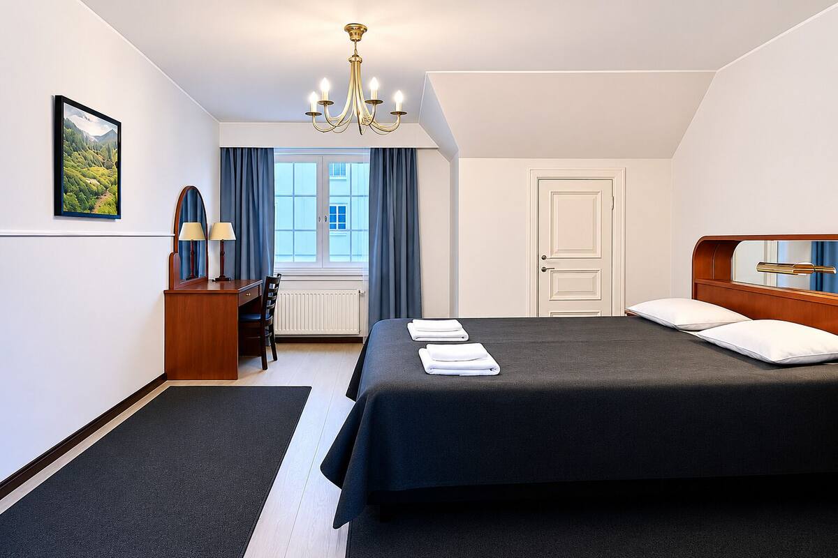 Ein Hotelzimmer mit einem großen Bett, einem Schreibtisch mit Stuhl, einer Schreibtischlampe, einem Fenster mit Vorhängen und einer Kristallleuchter.