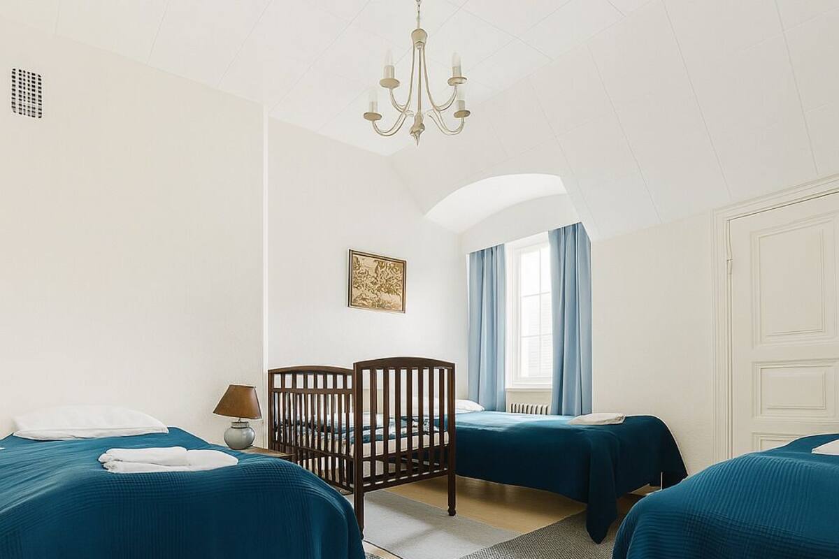 Ein Hotelzimmer mit zwei Betten, einem hölzernen Wiege, einer Kristallleuchter und einem Fenster mit Vorhängen.