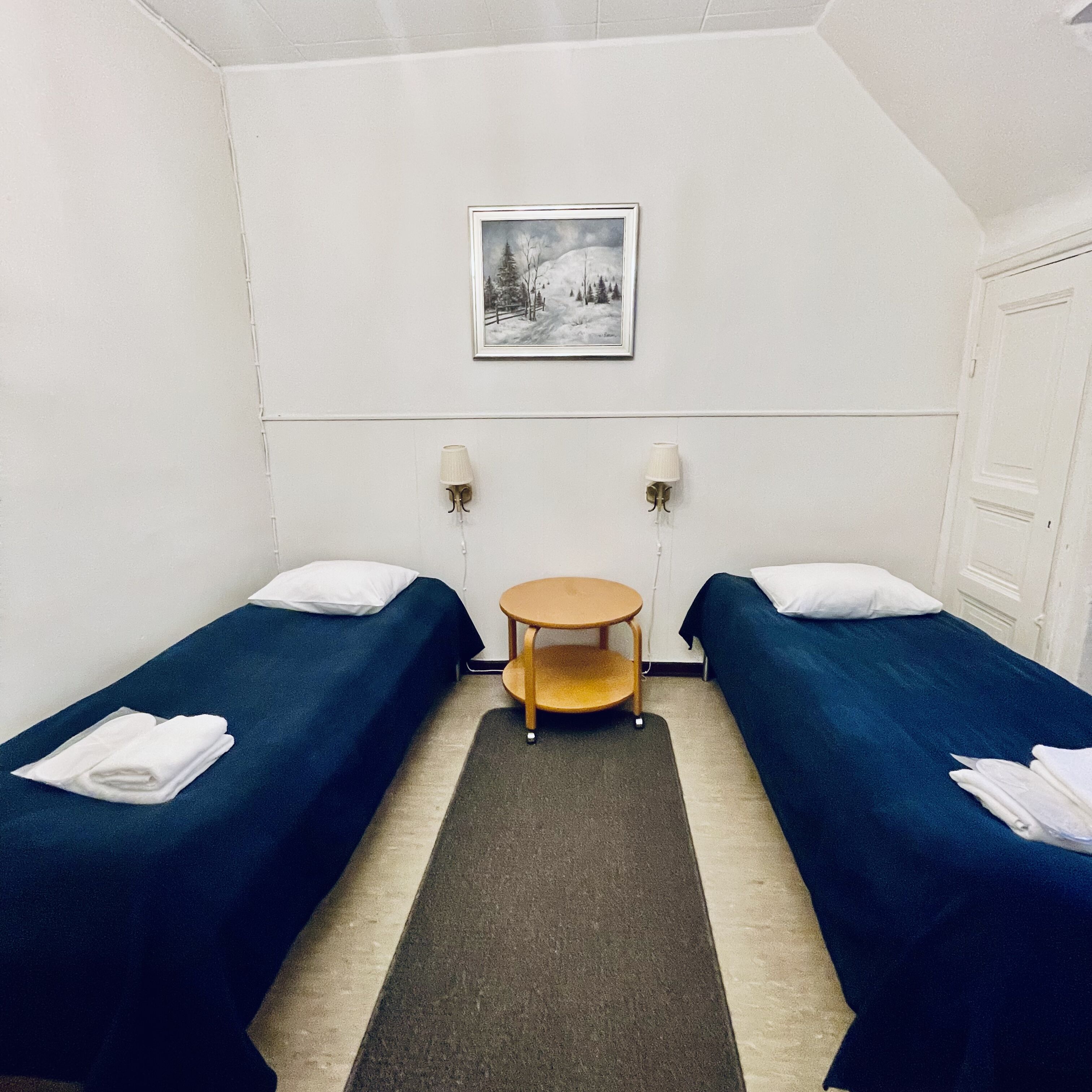 A small, simple room with two single beds, a small table, two wall lamps, and a framed picture on the wall.