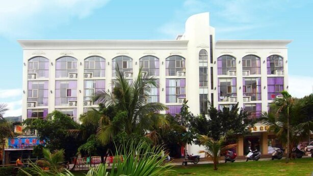 Un hotel de varios pisos con fachada blanca y detalles morados, rodeado de palmeras y vehículos estacionados.