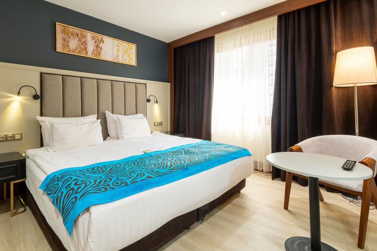 Habitación de hotel con cama, mesitas de noche, escritorio, silla, lámpara y ventana con cortinas.